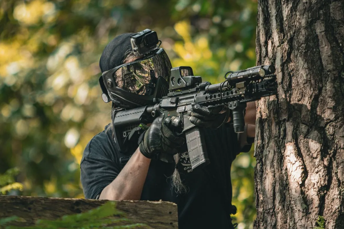 An airsoft player in a mask sheltering behind a tree in autumn light