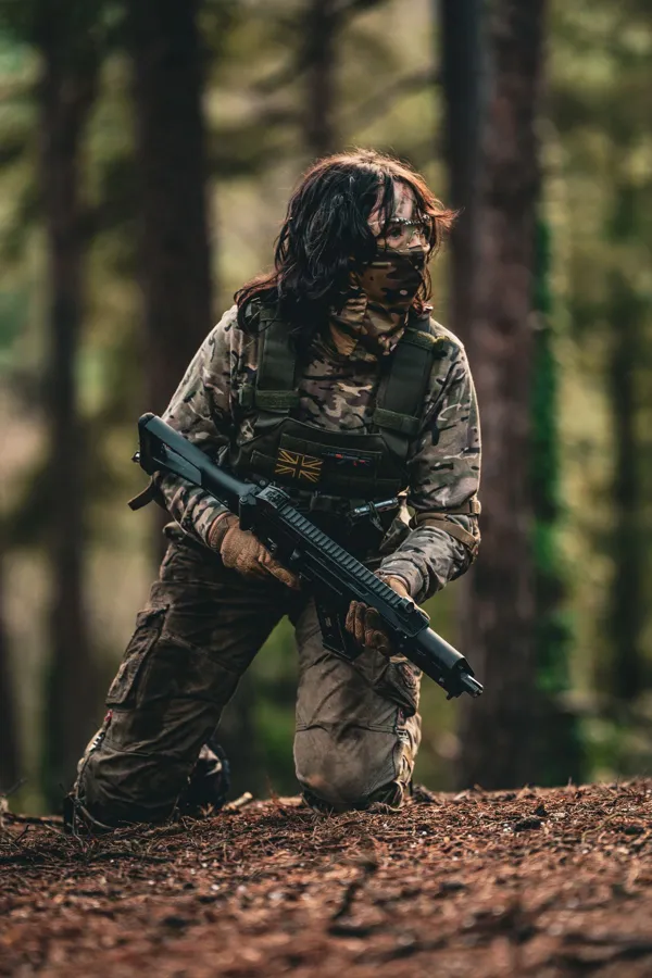 An airsoft player kneeling on a forest floor with a replica rifle