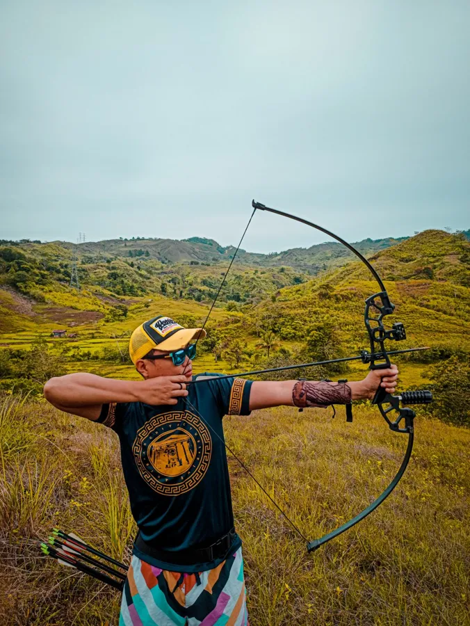 An archer in a bright shirt drawing a bow in grassy hills
