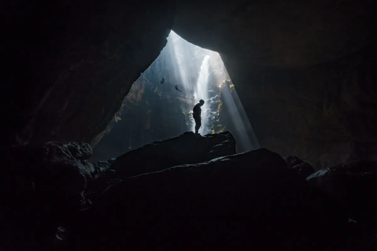Silhouette of a person exploring a vast cave with striking sunbeams filtering through the opening.