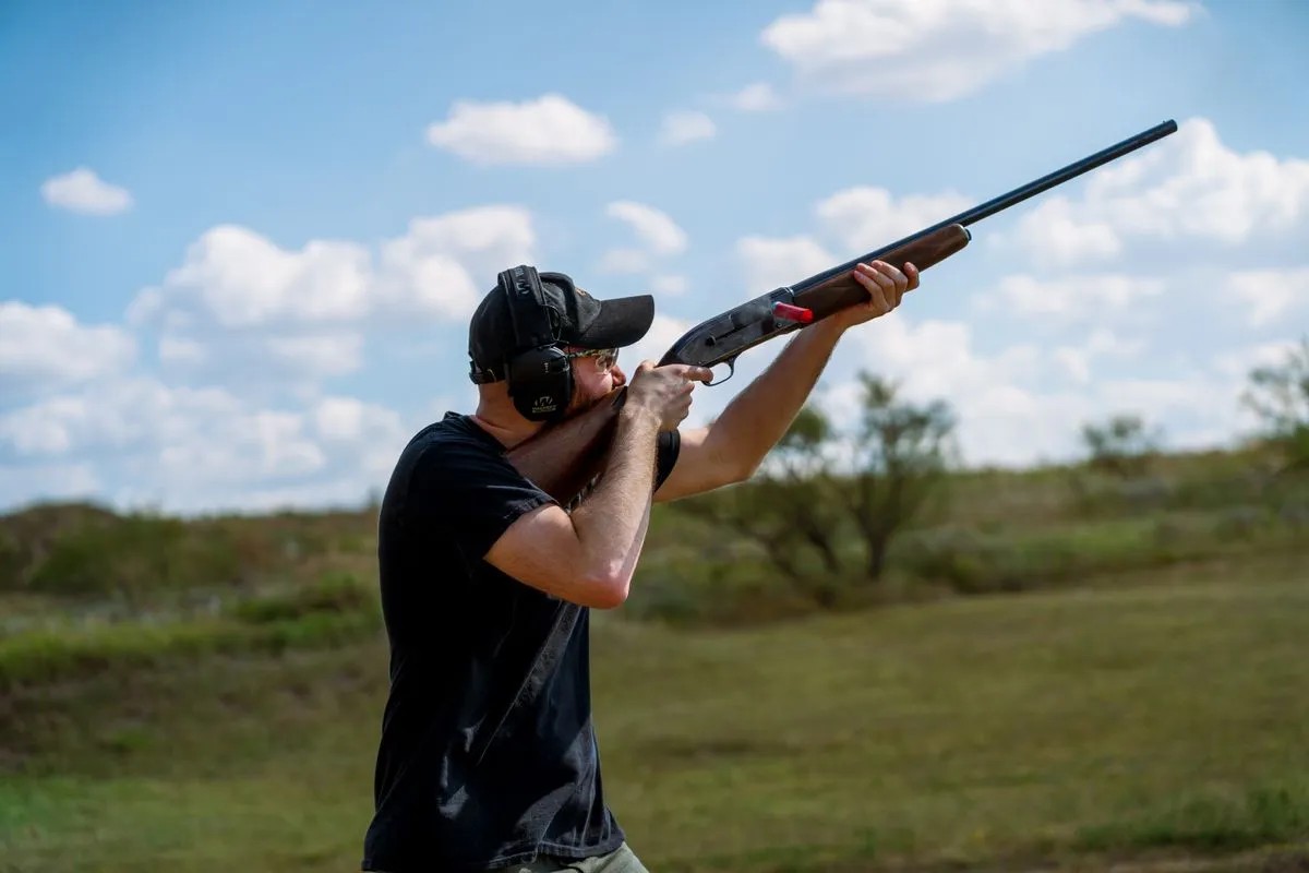 A man in ear defenders aiming a shotgun under a blue sky with clouds