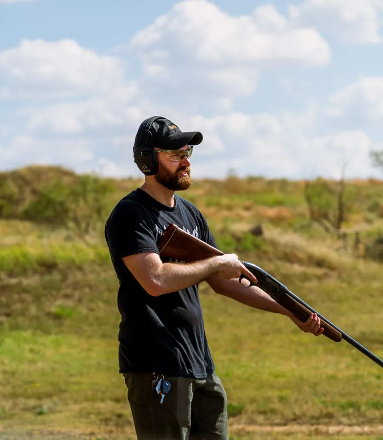 A man in ear defenders holding a shotgun in a grassy field