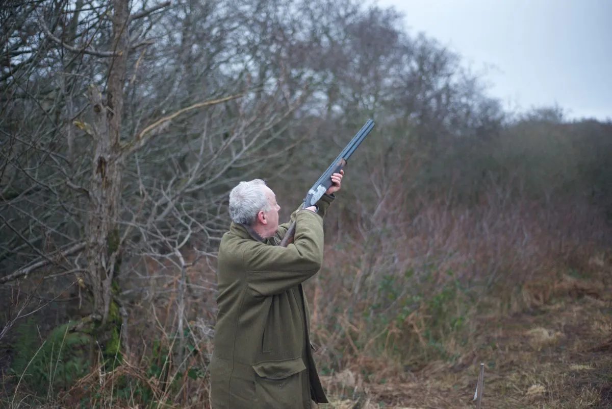 A shooter aiming a shotgun skyward at a clay target in a wooded setting