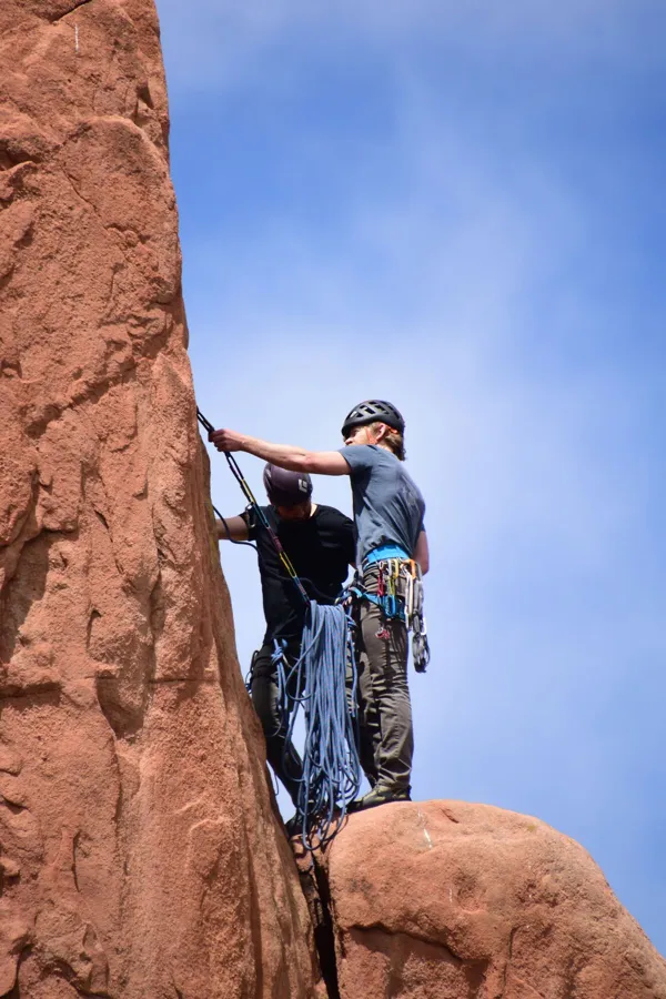 Two climbers with helmets and gear ascending a red sandstone cliff under a clear blue sky.