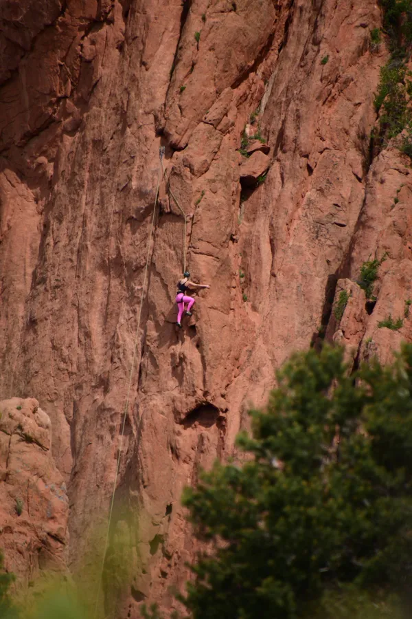 Adventurous climber scaling red rocky cliff with green foliage below.
