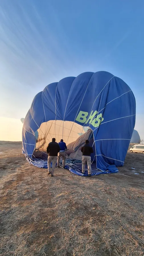 Three adults preparing a blue hot air balloon for takeoff during a clear sunrise.