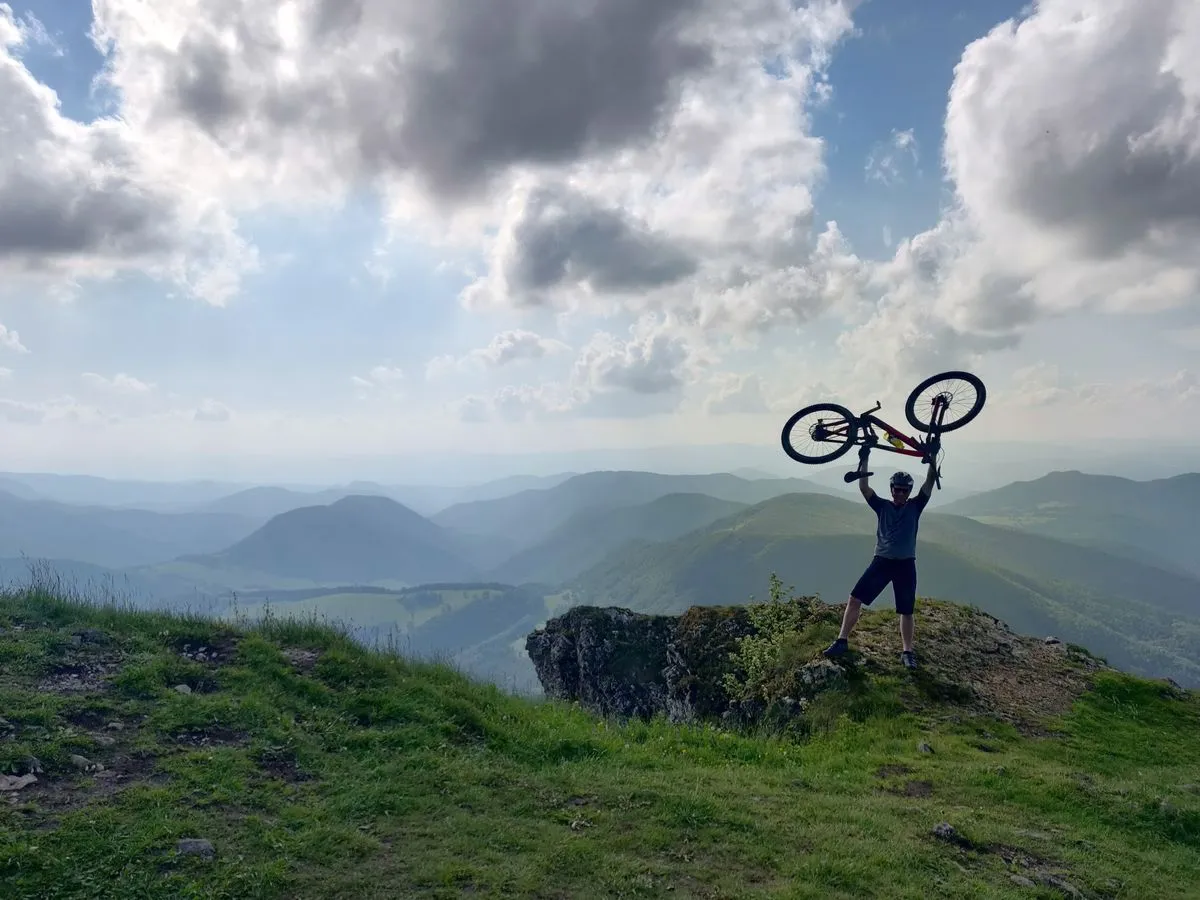 A rider holding a mountain bike overhead on a hilltop under a cloudy sky