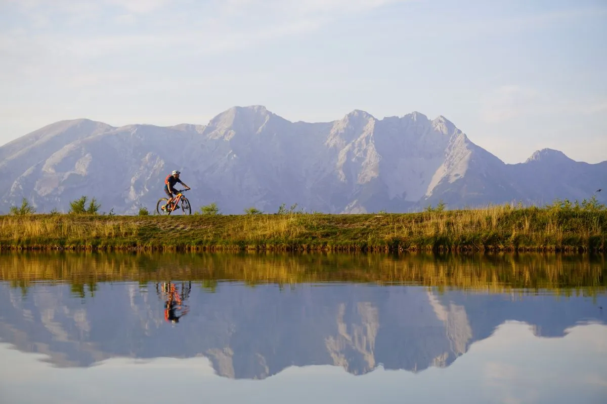 A rider on a path beside a still lake with mountains behind