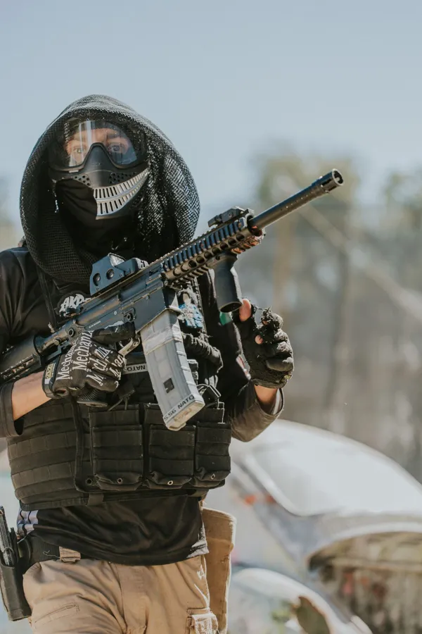 A paintball player in tactical gear and mask holding a paintball marker outdoors.