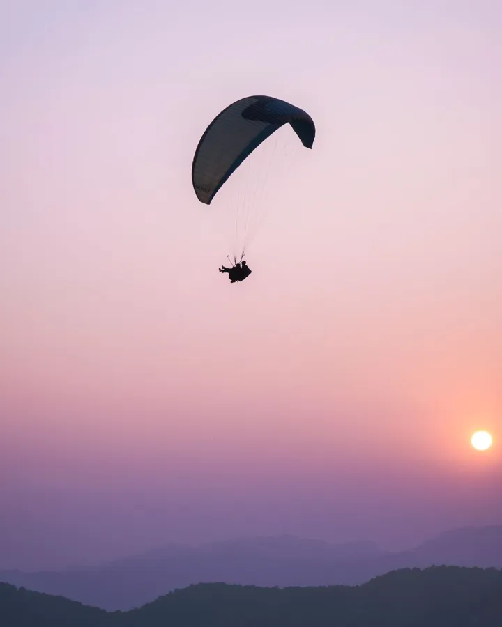 Silhouette of a paraglider soaring against a bright sunset over mountains.